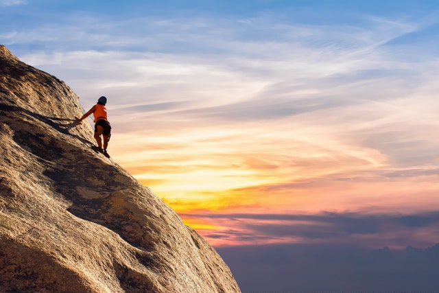 A hiker climbing a large rock in front of a sunset.